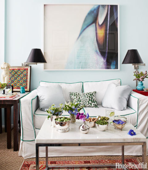 Petite living room with pale blue walls, bold art, and swing-arm sconces above the sofa. As seen in House Beautiful, designed by Nick Olsen,